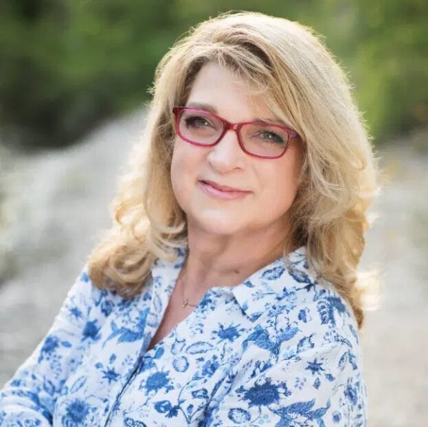 A woman with blonde hair and red glasses, wearing a blue-patterned shirt, stands outdoors with greenery in the background.